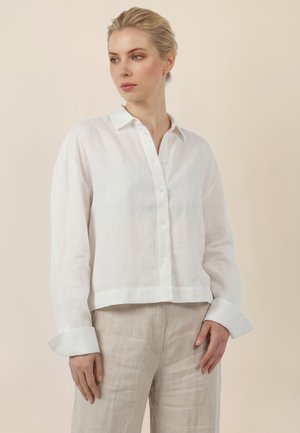 Frau mit blondem nach hinten gebundenem Haar, trägt ein weißes Hemd mit Knöpfen und beigen Hosen, schaut leicht nach links vor einem schlichten Hintergrund.