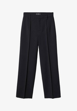 Femme portant un pull gris clair à manches longues et col en V, rentré dans un pantalon noir taille haute, les mains placées derrière son dos.