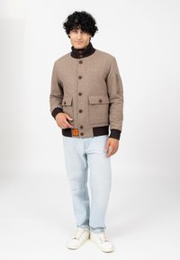 Mann mit lockigem Haar, der eine braune Jacke mit Knöpfen und großen Taschen, hellblaue Jeans und weiße Sneaker trägt, vor einem weißen Hintergrund steht.