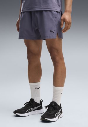 Personne portant un short violet, des chaussettes Puma blanches et des chaussures de course Puma noires, debout devant un fond uni en dégradé.