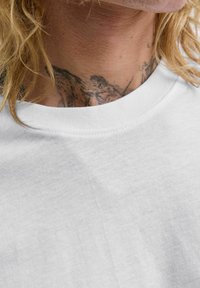 Artimas žmogaus kaklo vaizdas, kuriame matoma juoda tatuiruotė, dalinai atsiverianti virš baltos apvalios kaklo marškinėlės, ir šviesiaplaukės plaukų sruogos.