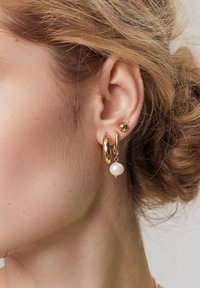 Złote kolczyki z fakturowanym kółkiem, gładką złotą kulką i wiszącą perłą. Projekt łączy różne kształty i wykończenia.