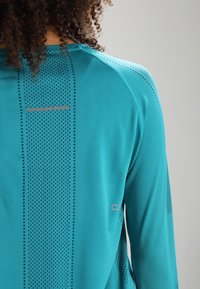 Langärmliges Sportshirt in Teal, mit einem strukturierten Muster auf der Rückseite und eleganten Designelementen aus dunkleren und reflektierenden Materialien.