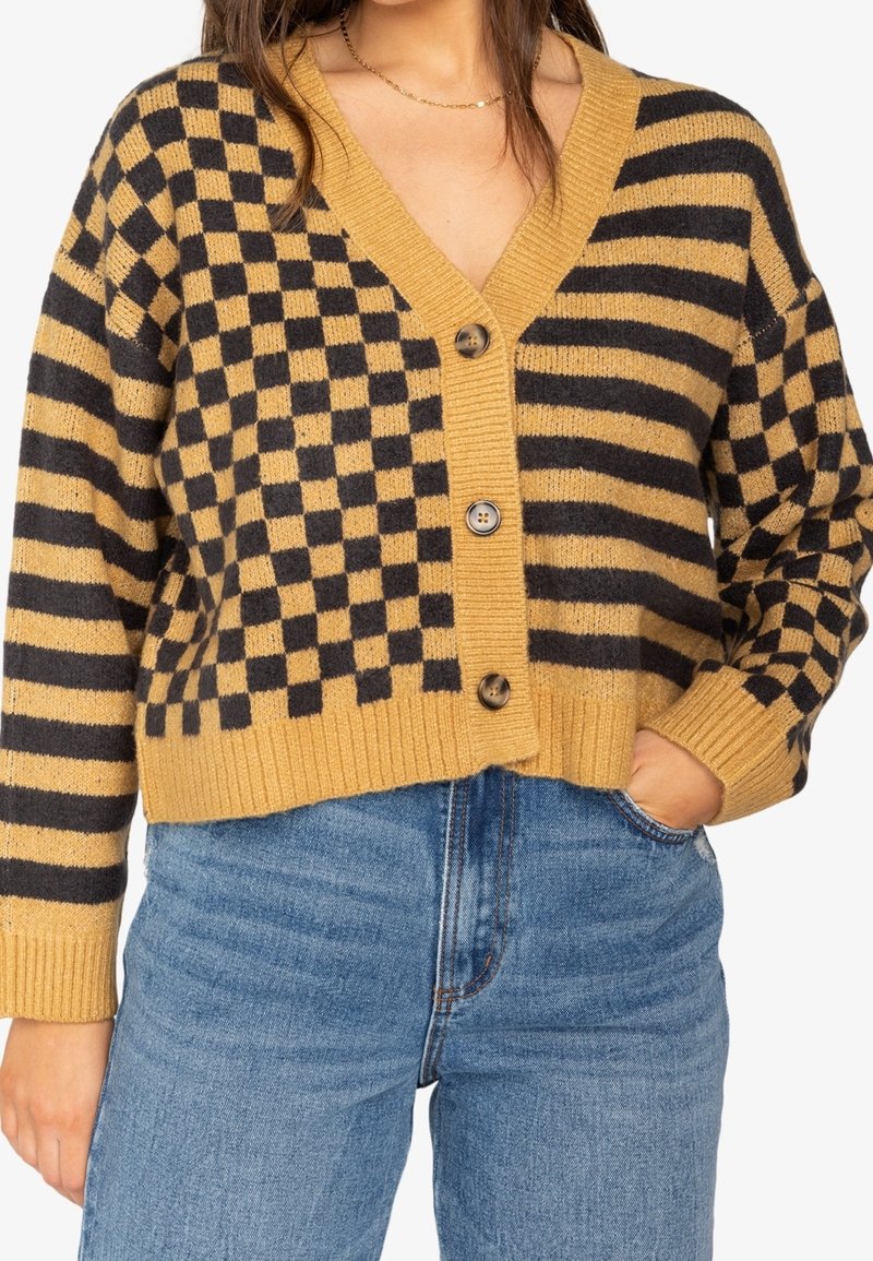 Femme portant un cardigan moutarde et noir boutonné avec des motifs damier et rayures, associé à un jean bleu taille haute.