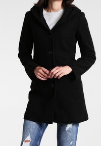 Abrigo de lana negro con capucha, con un cierre de botones en la parte delantera, dos bolsillos laterales y un diseño ajustado. Se lleva sobre una camiseta gris y jeans desgastados.