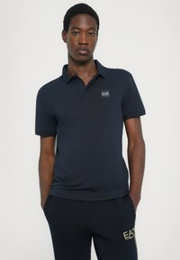 Polo azul-marinho com design de gola e mangas curtas. Apresenta um emblema quadrado no lado esquerdo do peito e um tecido suave e texturizado.