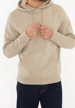 Homme portant un sweat à capuche beige et un jean bleu foncé tenant les lacets de la capuche avec les deux mains sur un fond blanc.