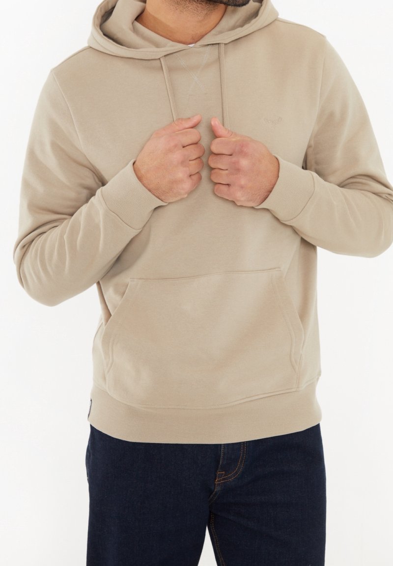 Homme portant un sweat à capuche beige et un jean bleu foncé tenant les lacets de la capuche avec les deux mains sur un fond blanc.