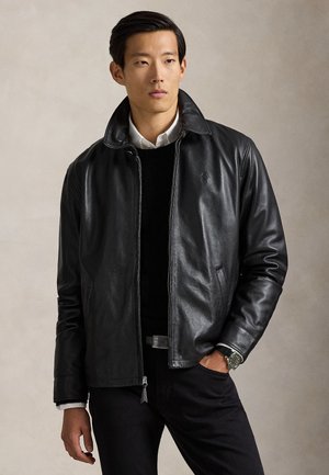 Mann trägt eine schwarze Lederjacke über einem schwarzen Pullover und einem weißen Hemd, mit einer Hand in der Tasche und einer Uhr am Handgelenk.