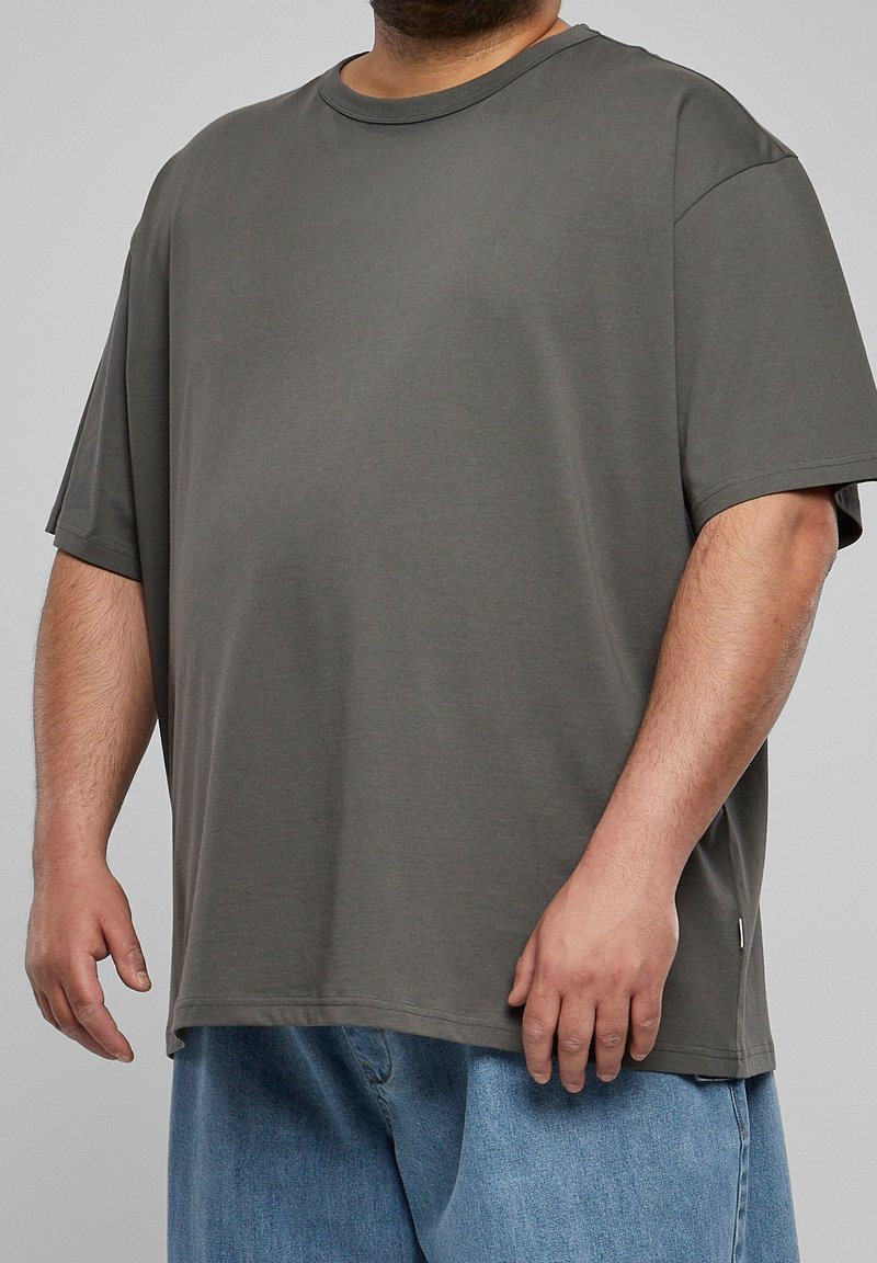 Homme portant un t-shirt gris à manches courtes ample et un jean bleu, se tenant devant un fond gris uni.