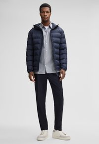 Marineblauwe donsjack met capuchon, grijs overhemd met knopen eronder, zwarte broek en beige sneakers. Stijlvolle textuur, modern ontwerp, slim fit.