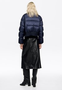 Granatowa kurtka puchowa o skróconym kroju, z prążkowanym wykończeniem, zestawiona z błyszczącą czarną skórzaną spódnicą oraz czarnymi, sięgającymi kolan, botkami na obcasie.
