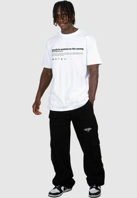 T-shirt blanc avec un texte noir indiquant "la jeunesse est gâchée sur les jeunes", associé à un pantalon cargo noir ample et des baskets avec des accents blancs.