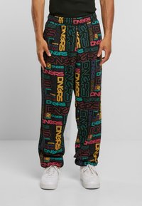 Mann som har på seg svarte bukser med multicolorede "DNGRS" tekstmønstre og hvite sneakers, stående foran en enkel grå bakgrunn.