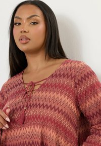 Blusa con motivo zigzag rosso e rosa, con scollatura a V, dettaglio con fiocco e frange, maniche lunghe e vestibilità rilassata. Tessuto testurizzato.