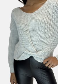 Lichtgrijze, gebreide trui met een diepe ronde halslijn en een gedraaide knoopdetail aan de taille, gecombineerd met glanzende zwarte leggings.