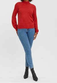 Roter Pullover mit langen Ärmeln und hohem Kragen, kombiniert mit blauen Skinny-Jeans und schwarzen Stiefeletten. Weiche Textur, taillierte Passform.