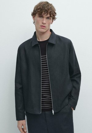 Jeune homme aux cheveux bouclés portant une veste zippée foncée par-dessus une chemise rayée noire et blanche, debout avec une main dans la poche devant un fond uni.