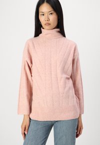 Sveternik s svetlo roza turtleneck iz ribanim robom in manšetami, ki vsebuje dvignjene vertikalne vzorce. Mehka tekstura, sproščen kroj, dolgi rokavi.