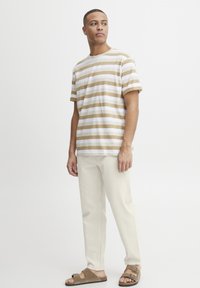 Stripet t-skjorte i beige og hvit, kombinert med kremfargede bukser. Modellen har på seg brune sandaler. Enkel design med korte ermer og rund hals.