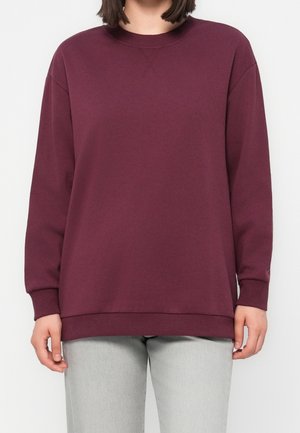 Femme portant un sweat-shirt bordeaux uni à manches longues avec col rond et un pantalon gris clair, debout sur un fond blanc.