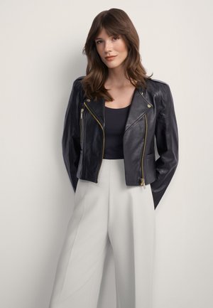 Młoda kobieta o brązowych włosach, ubrana w czarną skórzaną kurtkę, czarny top i białe, szerokie spodnie z wysokim stanem na jednolitym tle.