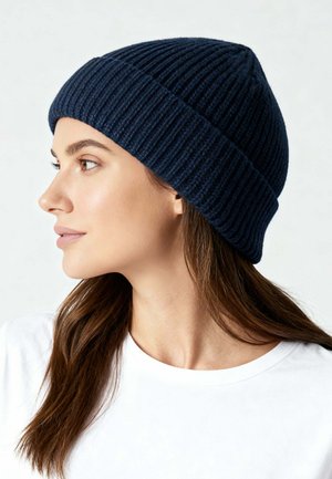 Vrouw met lang bruin haar die een marineblauwe geribbelde gebreide muts en een wit shirt met ronde hals draagt, kijkend naar links.