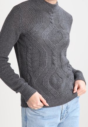 Pull gris foncé tricoté en câble avec des motifs tressés complexes, porté avec un jean bleu clair, mains posées négligemment dans les poches, debout devant un fond uni.