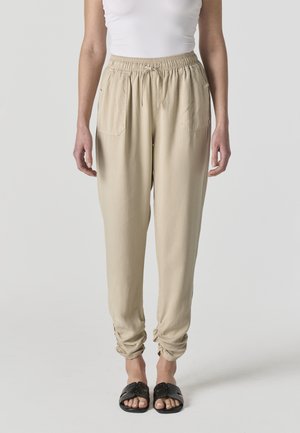 Mujer con pantalones jogger de cintura elástica beige, top sin mangas blanco y sandalias negras tipo slip-on, posando frente a un fondo claro y sencillo.