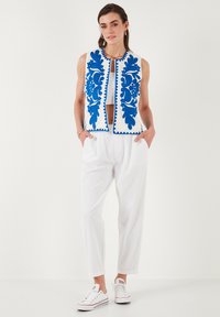 White sleeveless vest with blue floral embroidery, buttonless front closure, paired with white tapered pants and white sneakers.