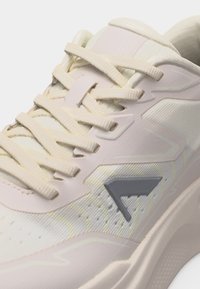 Κοντινό πλάνο ενός ανοιχτό μπεζ αθλητικού παπουτσιού με υφασμάτινη υφή, κρεμ κορδόνια και σκούρο γκρι λογότυπο στη πλευρά κοντά στη σόλα.