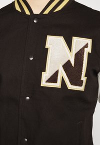 Czarna kurtka varsity z beżowym kołnierzem w paski oraz dużą naszywką z literą "N" w kolorze beżowym i ciemnobrązowym na piersi.