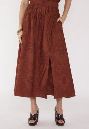 Femme portant une jupe midi à motifs couleur rouille avec une fente avant, associée à des sandales noires à talons hauts et des bracelets dorés au poignet gauche.