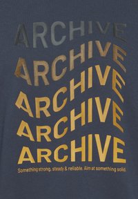 Camiseta azul marino con la palabra "ARCHIVE" en diferentes tamaños de fuente y tonos dorados, creando un diseño curvado. El texto incluye un eslogan en la parte inferior.
