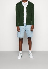 Groene gebreide cardigan met bruine knopen, wit T-shirt, lichtblauwe denim shorts met rafelige randen, en witte sneakers met groene strepen.