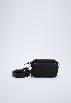 Sort rektangulær crossbody taske med to lynlåskompartementer og en aftagelig skulderrem på en hvid baggrund.