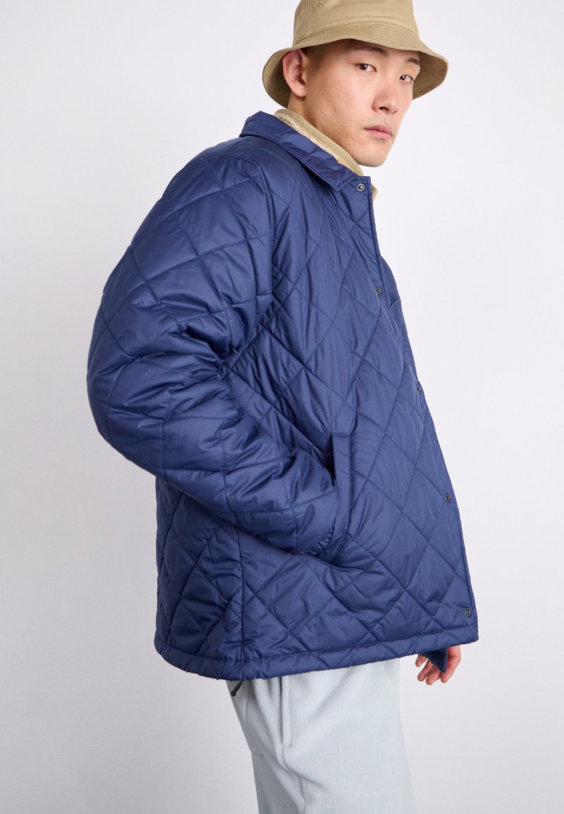 Veste matelassée bleu marine avec boutons-pression et poches latérales. Comprend un col et une coupe décontractée. Le tissu semble lisse et isolant.