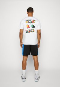 Vit t-shirt med färggrafiska, lekfulla text och former på baksidan, i kombination med svarta shorts med blå sidostripes.