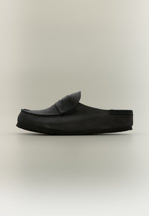 Zapato mule negro sin cordones con parte superior de ante y detalle de tira de cuero, suela plana y talón abierto, mostrado sobre un fondo claro y sencillo.