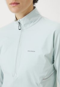 Nærbilde av en person som har på seg en lys blå Salomon zip-up jakke med høy krage, som viser merkevarens logo på brystet.