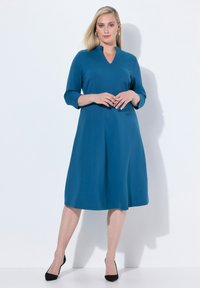 Turkusowa sukienka z dekoltem w serek, rękawami o długości trzy czwarte i luźnym fasonem, sięgająca poniżej kolan. Założona z czarnymi szpilkami. Gładka faktura.