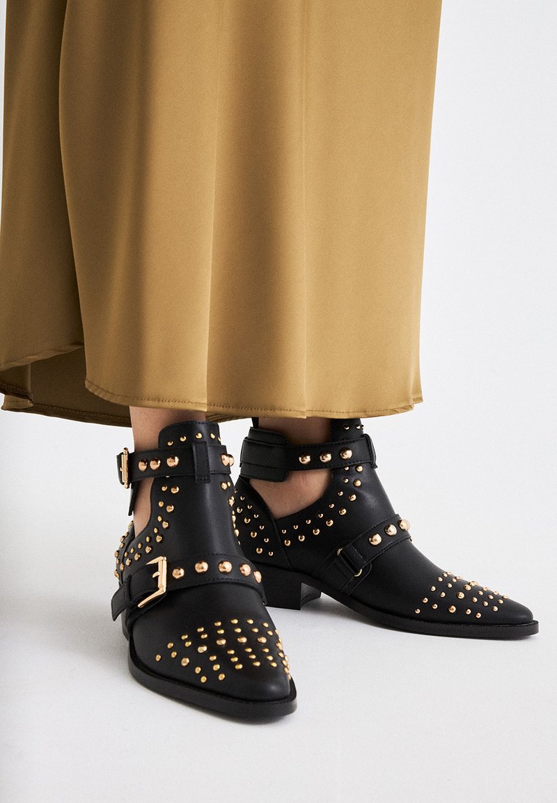 Botas de tobillo de cuero negro con tachuelas doradas, punta afilada, hebillas y tacón bajo, combinadas con una falda larga beige.