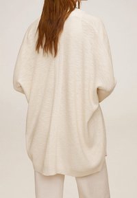Gilet oversized de couleur crème en doux tissu tricoté texturé. Présente des épaules tombantes et une coupe décontractée, avec un détail de couture au centre.