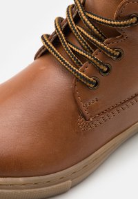 Bottine en cuir marron avec des lacets ronds noirs et bruns, œillets en métal et semelle texturée avec des détails de couture sur le côté.