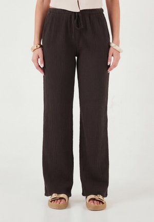 Nő, aki laza, sötétbarna, texturált nadrágot visel megkötött derékpánttal, bézs szandált, krémszínű felsőt és karkötőket mindkét csuklóján.