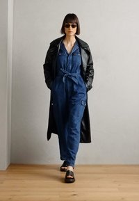 Tuta in denim con maniche lunghe, vestibilità ampia e cintura da annodare. Abbinata a un trench coat in pelle nera e sandali neri con punta aperta.