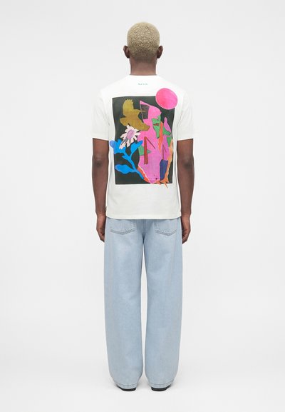 Άτομο με κοντά ξανθά μαλλιά φοράει λευκό μπλουζάκι με πολύχρωμο αφηρημένο σχέδιο και ανοιχτά μπλε παντελόνια φαρδιάς γραμμής, στέκεται γυρισμένο προς τα πίσω.