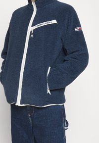 Námořnická modrá fleecová bunda s bílým zipem a podšívkou. Na hrudi má logo "TOMMY JEANS" a na rukávu malou vlajkovou akcentaci.