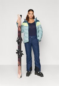 Pufi zöld sídzseki sárga kiegészítőkkel, tengerészkék nadrág és fekete sícipő. Közben egy barna sílécet tart fekete rögzítőkkel és piros felirattal.