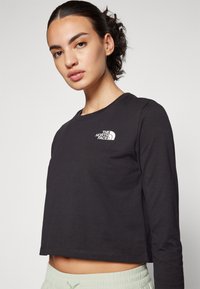Fekete, hosszú ujjú crop top, pamutból készült, fehér logóval a mellkasán, kerek nyakkivágással és laza illeszkedéssel.